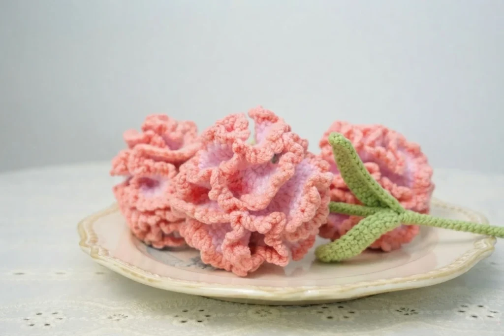 Three pink ruffled crochet carnations with green stems and leaves resting on a decorative plate on a lace tablecloth