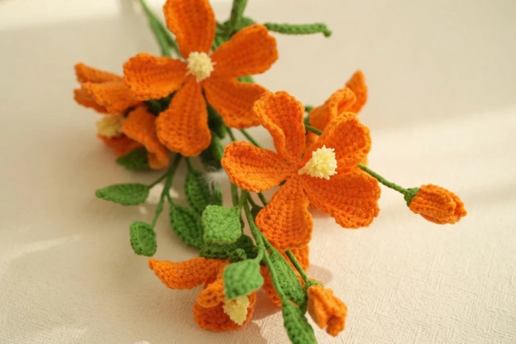 Orange crochet flowers with pale yellow centers, green leaves and small buds lying on a light surface