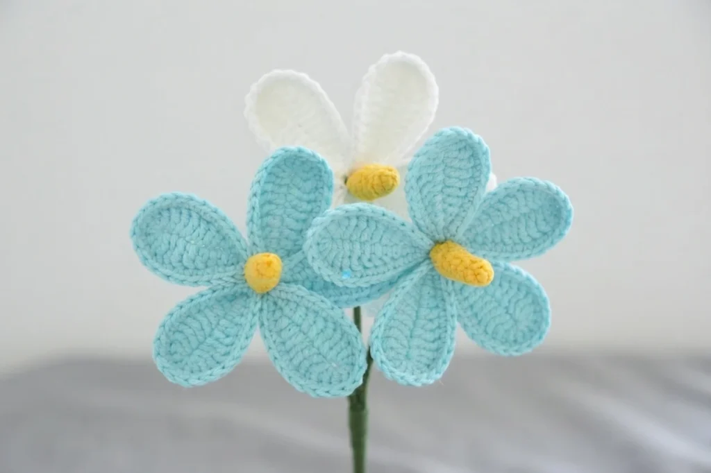Three crocheted flowers with light blue and white oval petals and yellow centers on a green stem