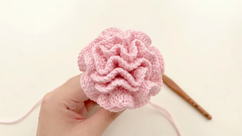 Hand holding a pale pink crocheted carnation with ruffled petals, with a wooden crochet hook in the background