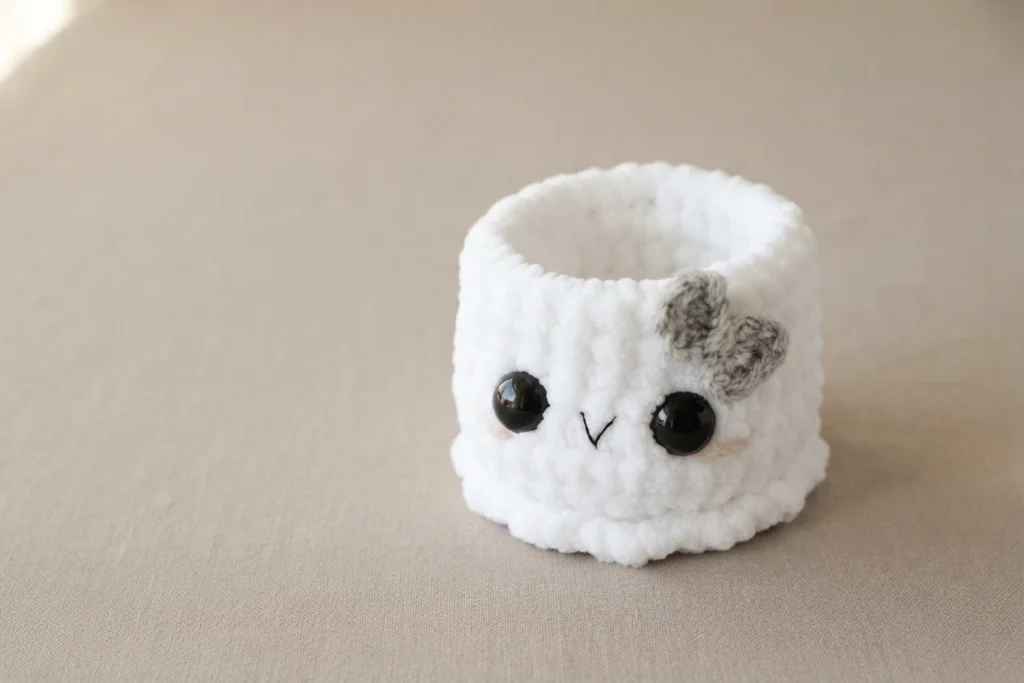 Single white crochet ghost gift holder with two black safety eyes, an embroidered smile, a small grey yarn bow on the right side of its head and a scalloped bottom edge, resting empty on a beige fabric surface.