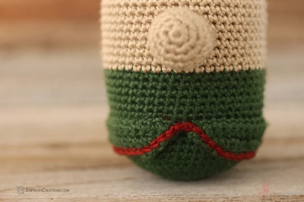 Crocheted elf gnome body on a wooden surface, with a cream colored upper section, a green tunic below and a red slip stitch trim forming a shallow V along the lower edge.
