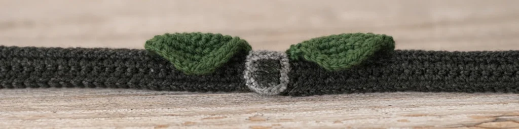 Crocheted dark charcoal belt laid flat on a light wood surface, with two green collars worked on either side of a grey buckle.