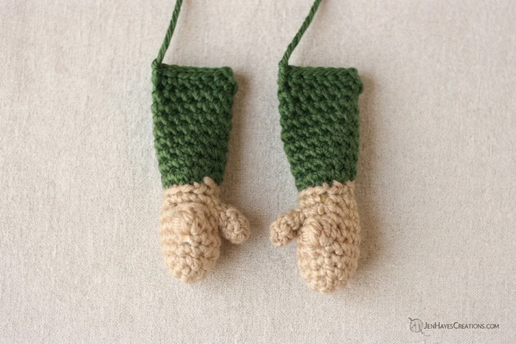 Two crocheted elf gnome arms laid side by side on a light textured surface, each with a tan stuffed hand and a dark green sleeve, a long green yarn tail at the top of each arm and a small designer watermark in the lower right corner.