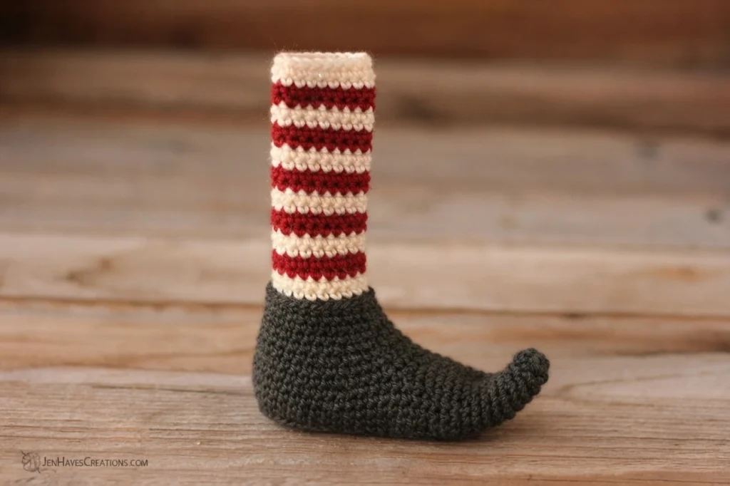Single crochet elf shoe with a charcoal grey foot and a red and white striped leg, standing upright on a wooden surface, with a JenHayesCreations.com watermark in the lower left corner.