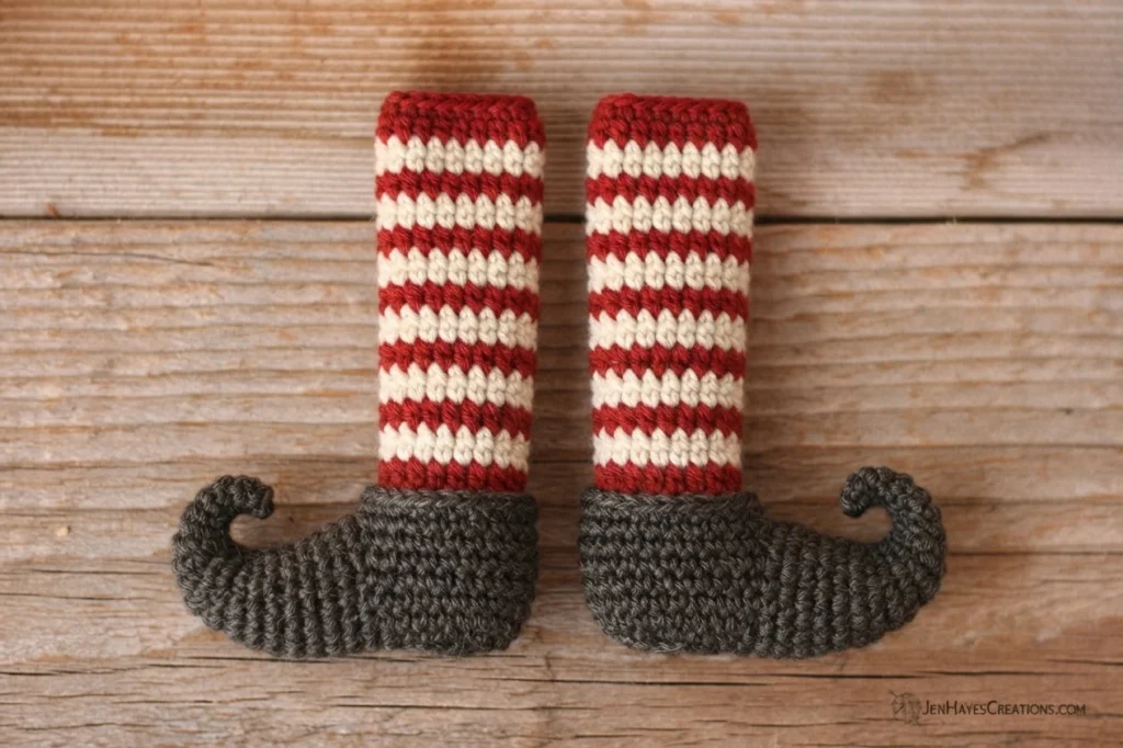 Two crocheted elf gnome legs lying side by side on a wooden surface, each with a red and cream striped stocking and a dark grey curled-toe shoe.