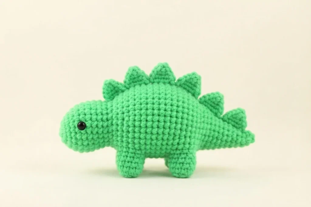 Green crochet dinosaur amigurumi with pointed spikes along its back, four stubby legs, one black safety eye and a small tail, photographed on a plain light background.