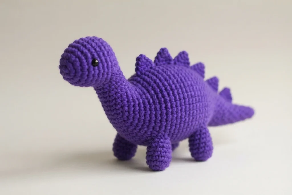 Finished purple crochet dinosaur amigurumi with spikes along its back, stubby legs and a black safety eye, standing in side view on a light background.
