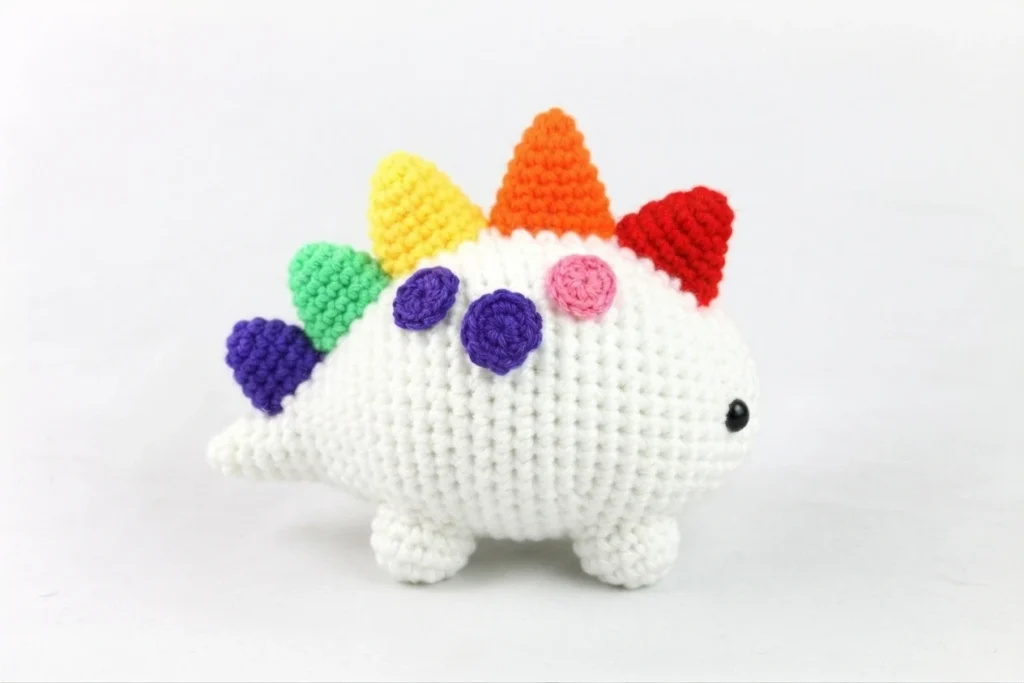 White crocheted dinosaur amigurumi with rainbow spikes along its back, small colored spots, black safety eyes and stubby legs on a plain white background.