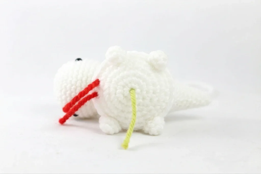 White crochet dinosaur amigurumi in progress with stubby legs, safety eyes, red stitch markers and loose yarn tails