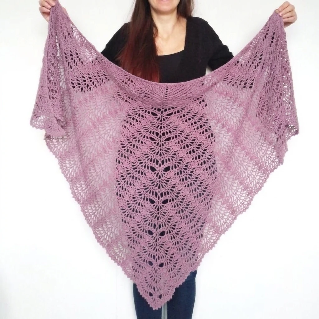 Person in a black top holding up a triangular pink crochet lace shawl with fan motifs, shown against a plain white wall.