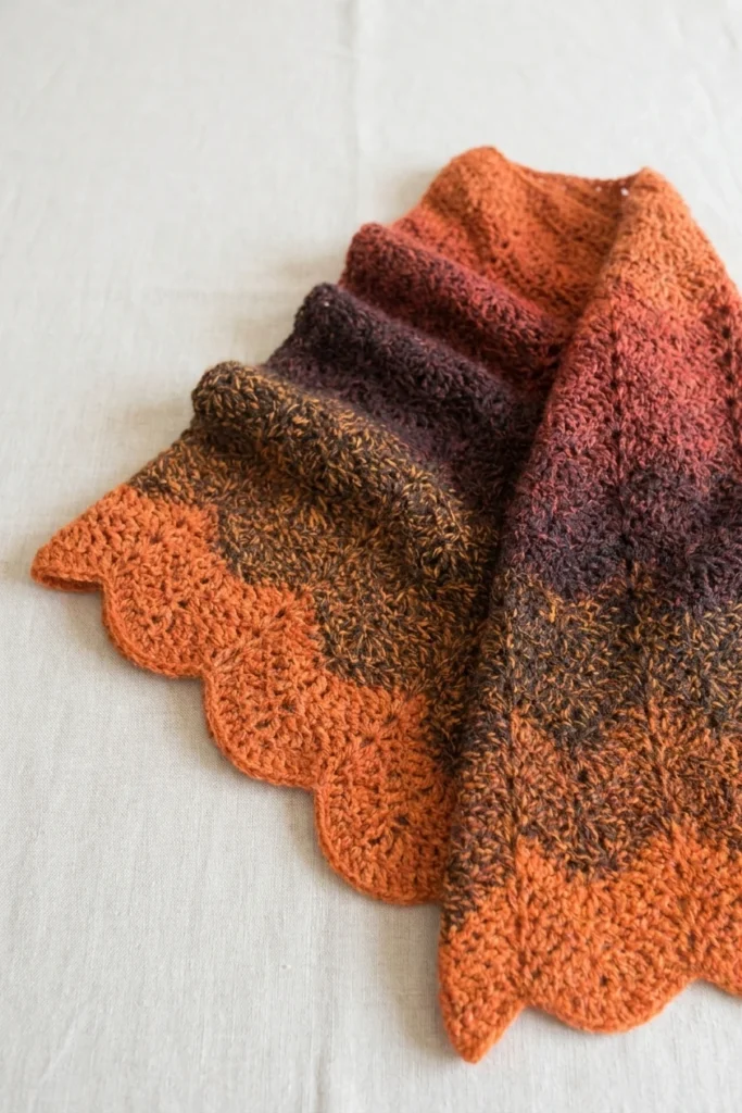 Crocheted ripple scarf with scalloped edges in orange, rust and brown gradient yarn, folded and laid flat on a light linen surface.