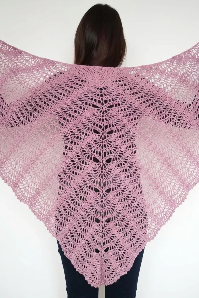 A person seen from behind wearing a pink triangular crochet shawl with open lace fan motifs draped over the shoulders, shown against a plain light background with dark trousers visible below.
