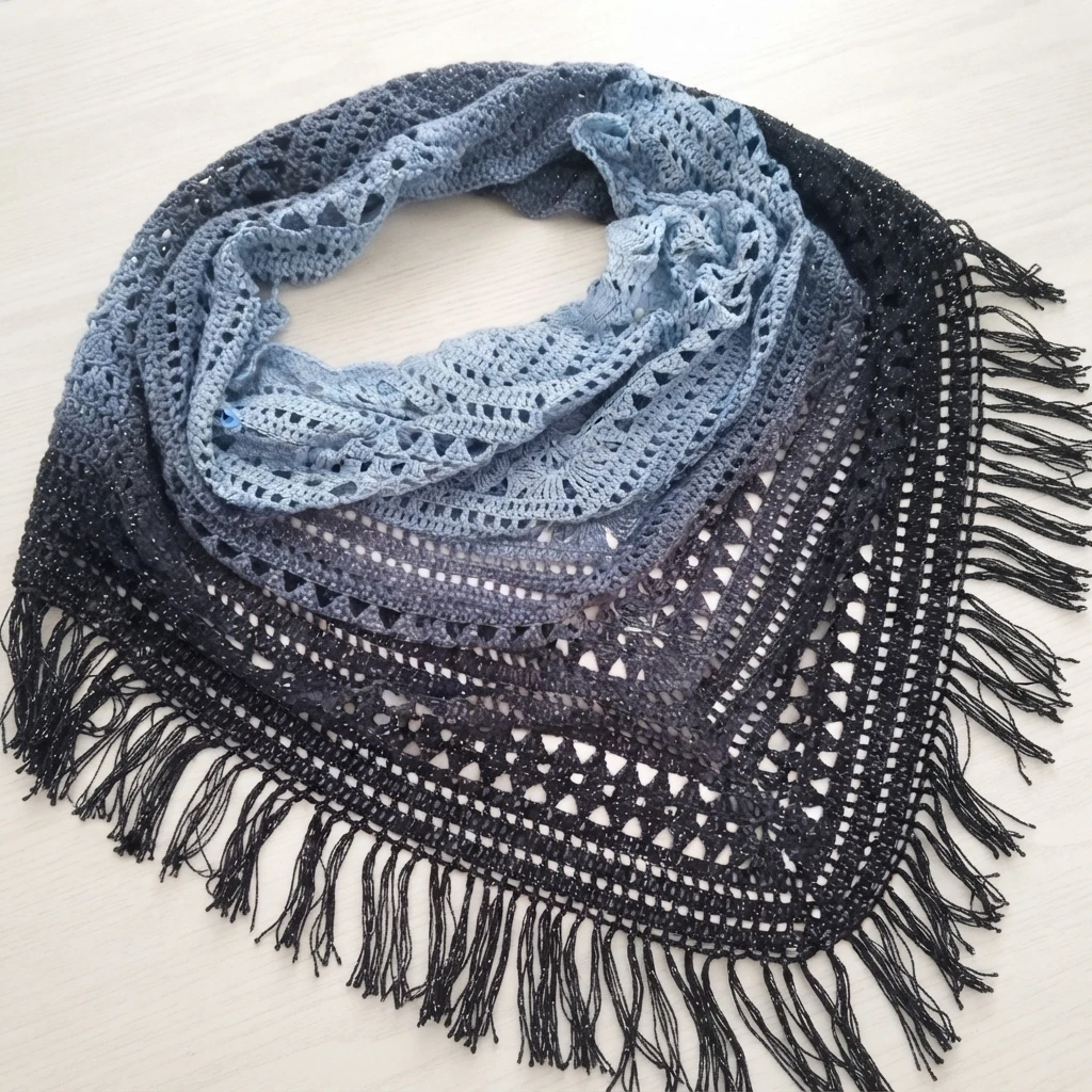 Blue to black gradient triangular crochet shawl with an openwork lace border and long fringe, laid flat on a light wood surface