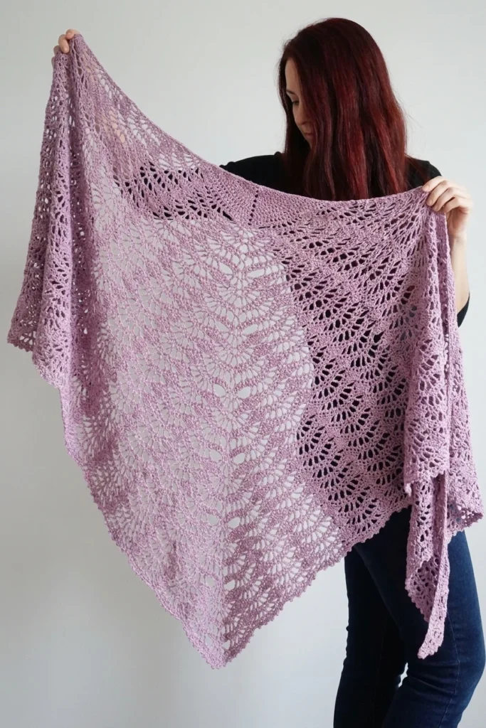 Woman with long dark red hair holding up a large lilac triangular lace shawl with open fan motifs and a scalloped edge, standing against a plain white wall in a black top and dark jeans.