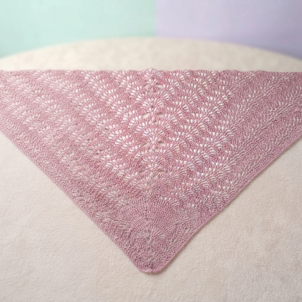 Pink triangular crochet lace shawl with open fan motifs, laid flat on a pale pastel surface