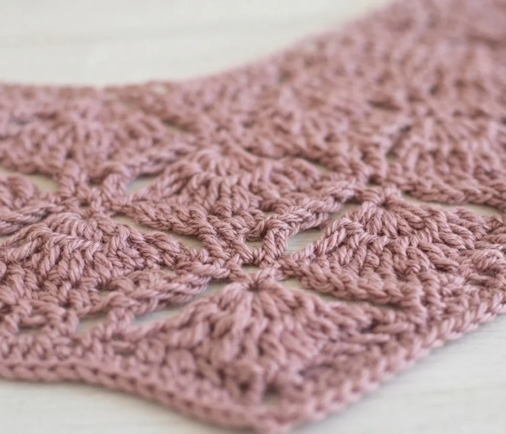 Close-up of dusty pink crochet lace fabric with repeating openwork fan motifs worked in rows, resting on a pale wooden surface.