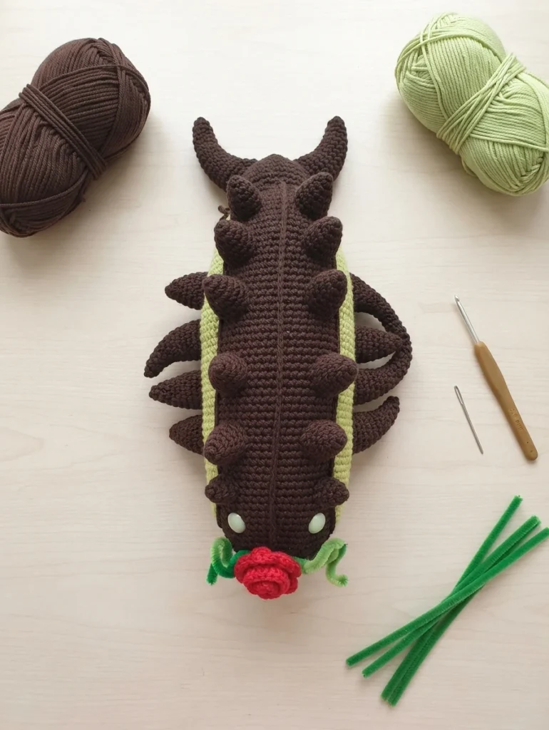 Flat lay of a crocheted brown and light green worm amigurumi with rows of brown spikes along its back and small pale eyes, a small red crocheted rose at its head, arranged on a pale wood surface next to a ball of brown yarn, a ball of light