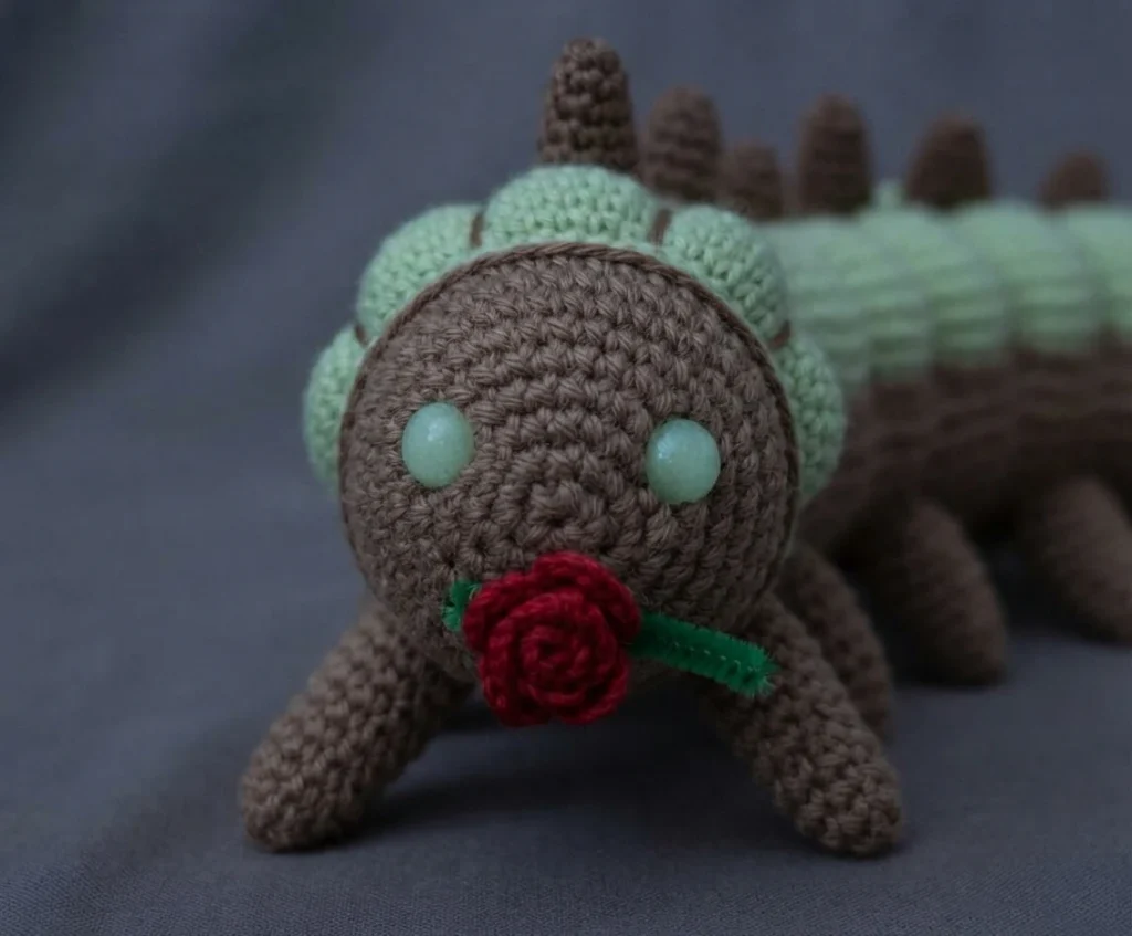 Crocheted worm amigurumi with a brown head and light green banded body, brown spikes along the back and tail, pale green round eyes and a small red crocheted rose with a green pipe cleaner stem near its mouth, resting on gray fabric.
