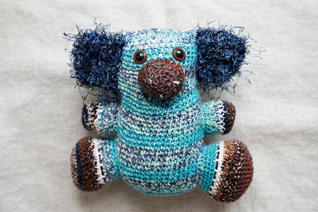 Crocheted amigurumi koala in striped turquoise yarn with fluffy dark blue ears, a brown nose and brown safety eyes, seated on a neutral fabric background.