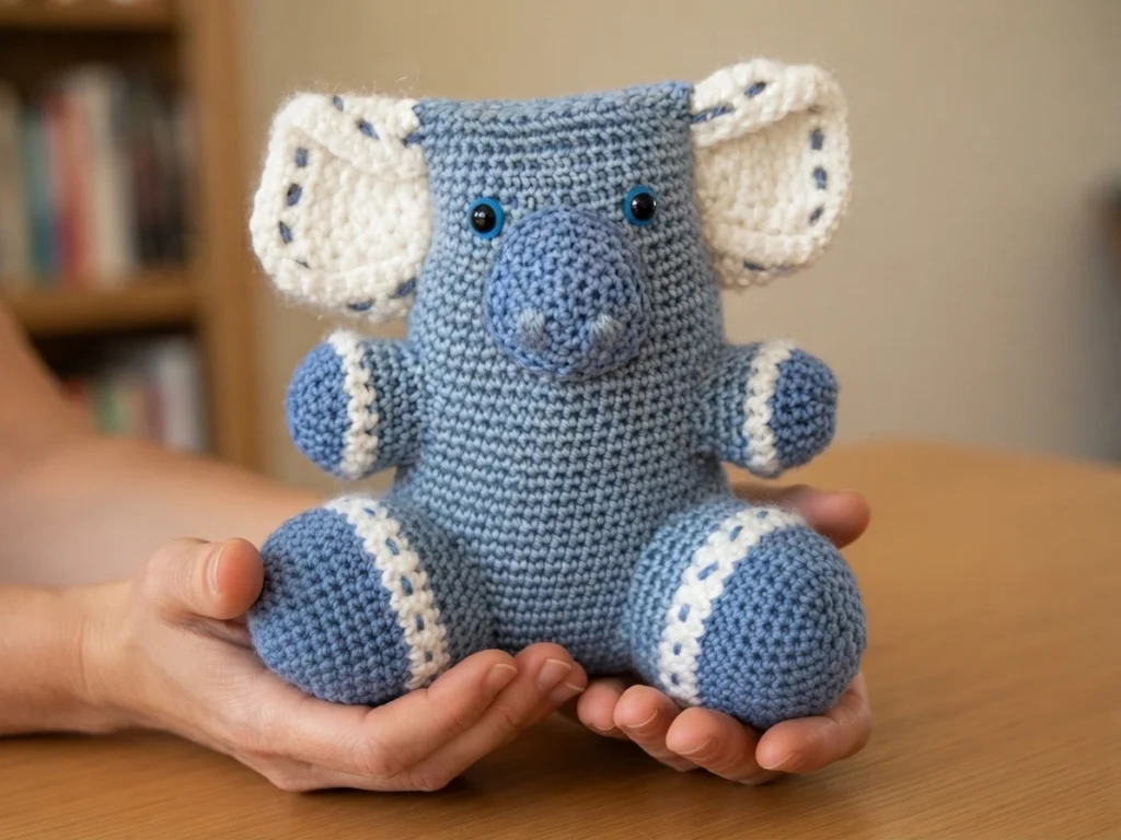 Crocheted blue koala toy with white ears and white-trimmed limbs, held in two hands on a wooden table.