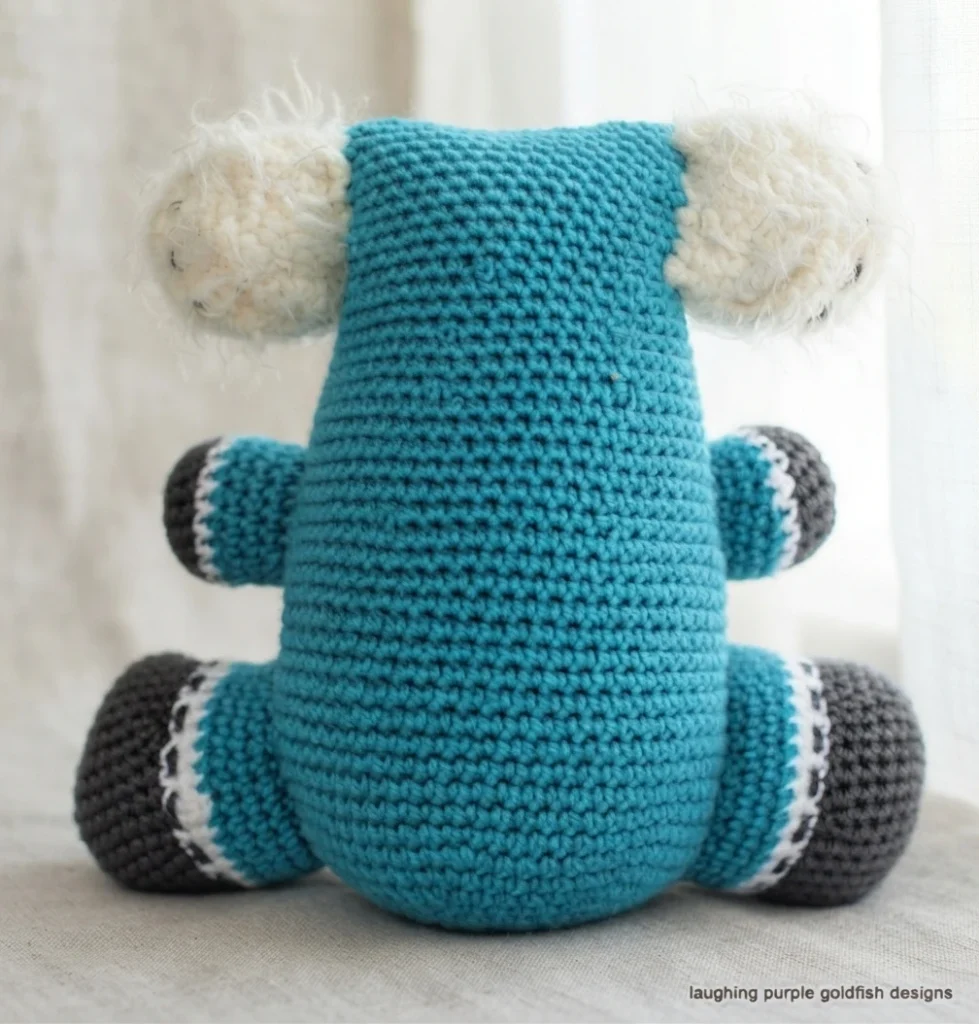Back view of a blue crocheted koala toy with fluffy white ears, dark gray paws with white trim, sitting on a light surface.