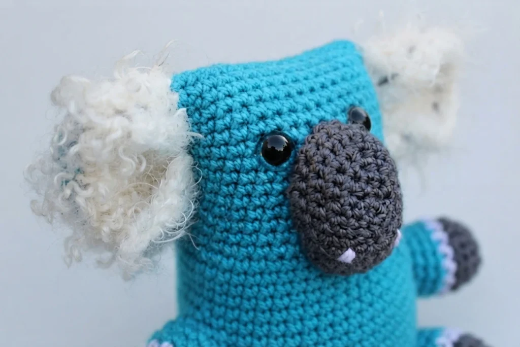 Crocheted blue koala amigurumi with fluffy white inner ears, dark gray muzzle and paw tips, and glossy black eyes.