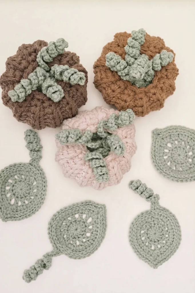 Three crocheted pumpkins with sage-green curly stems and several matching leaf-shaped coasters on a light background.
