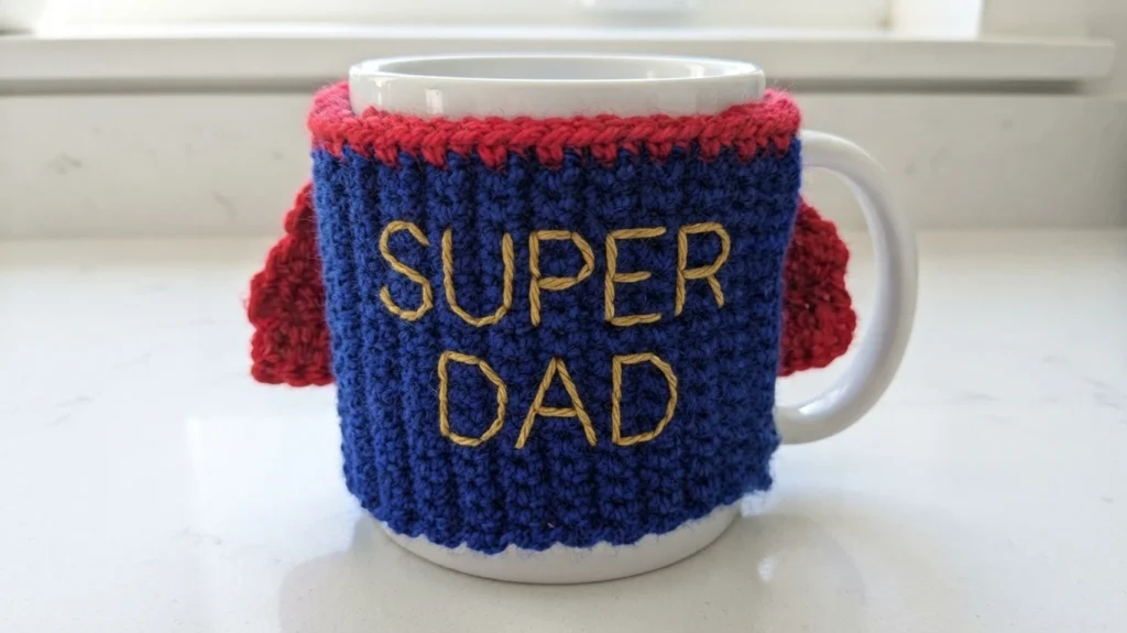 White mug with a blue crocheted cosy with red top trim and small red cape, embroidered with yellow SUPER DAD lettering