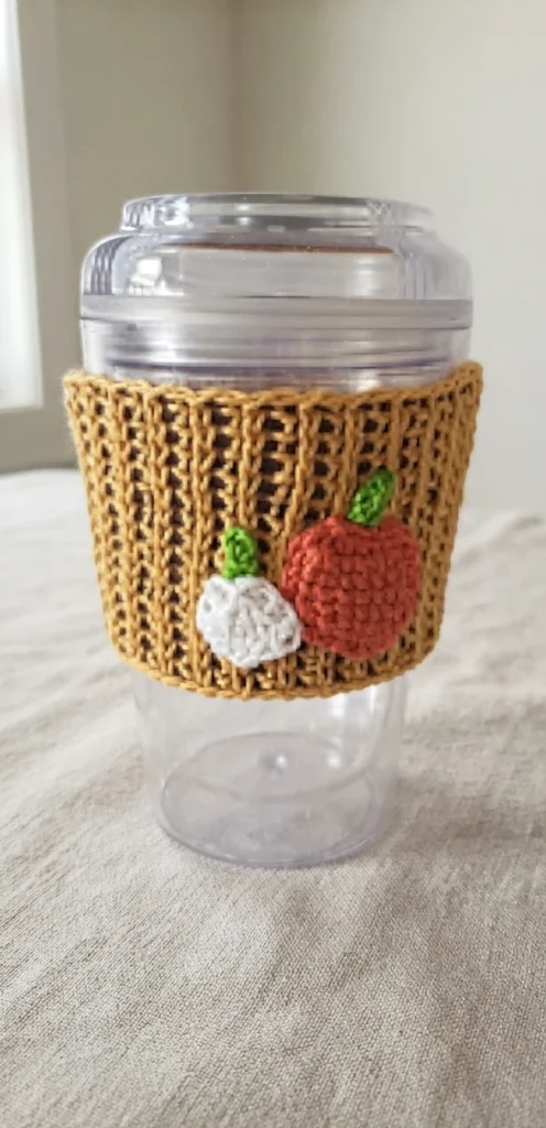 Crocheted mustard ribbed cup cozy wrapped around a clear plastic cup with a lid, with a white pumpkin motif and an orange pumpkin motif with green stems attached to the front.