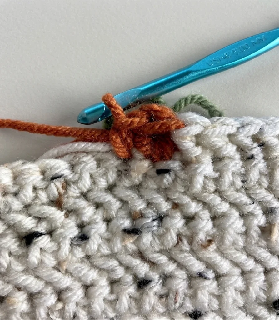 Gros plan d’un crochet bleu travaillant du fil orange sur un tissu crocheté ivoire chiné.