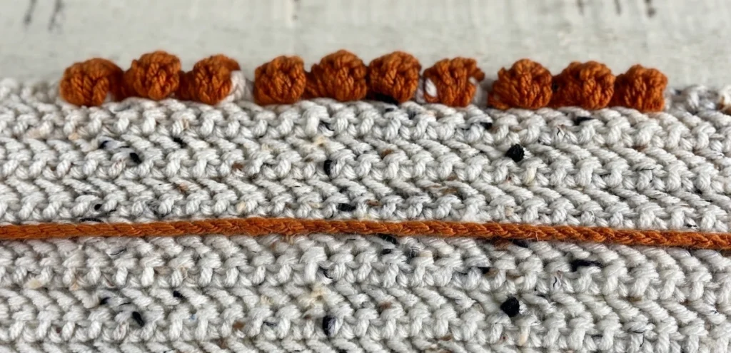 Close-up of raised orange crochet stitches on flecked ivory fabric, with an orange strand running horizontally across it.