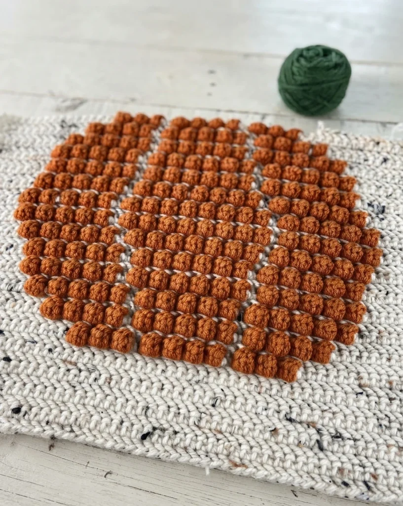 Panneau crocheté écru moucheté avec un motif orange en relief et une pelote de laine verte posée au-dessus.