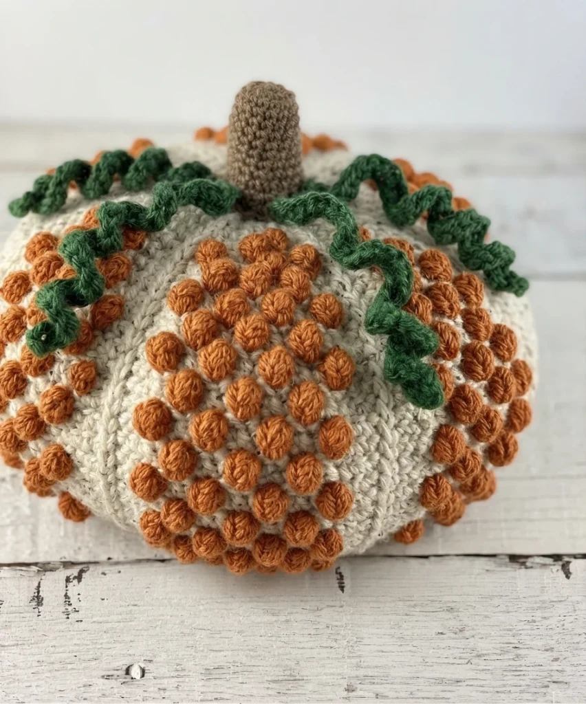 A round cream crocheted pumpkin with raised orange bobbles, a brown stem and curled green vines on a whitewashed wooden surface.