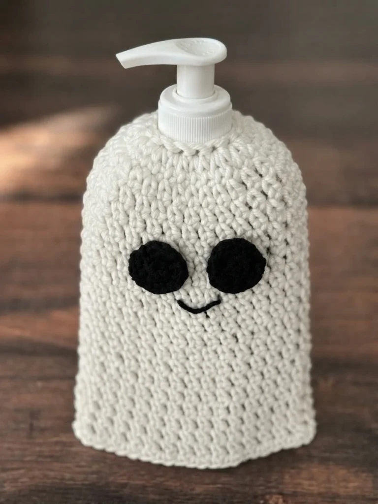 White crocheted ghost soap cover with two black crocheted eyes and an embroidered black smile, fitted over a white pump bottle and photographed on a wooden surface.