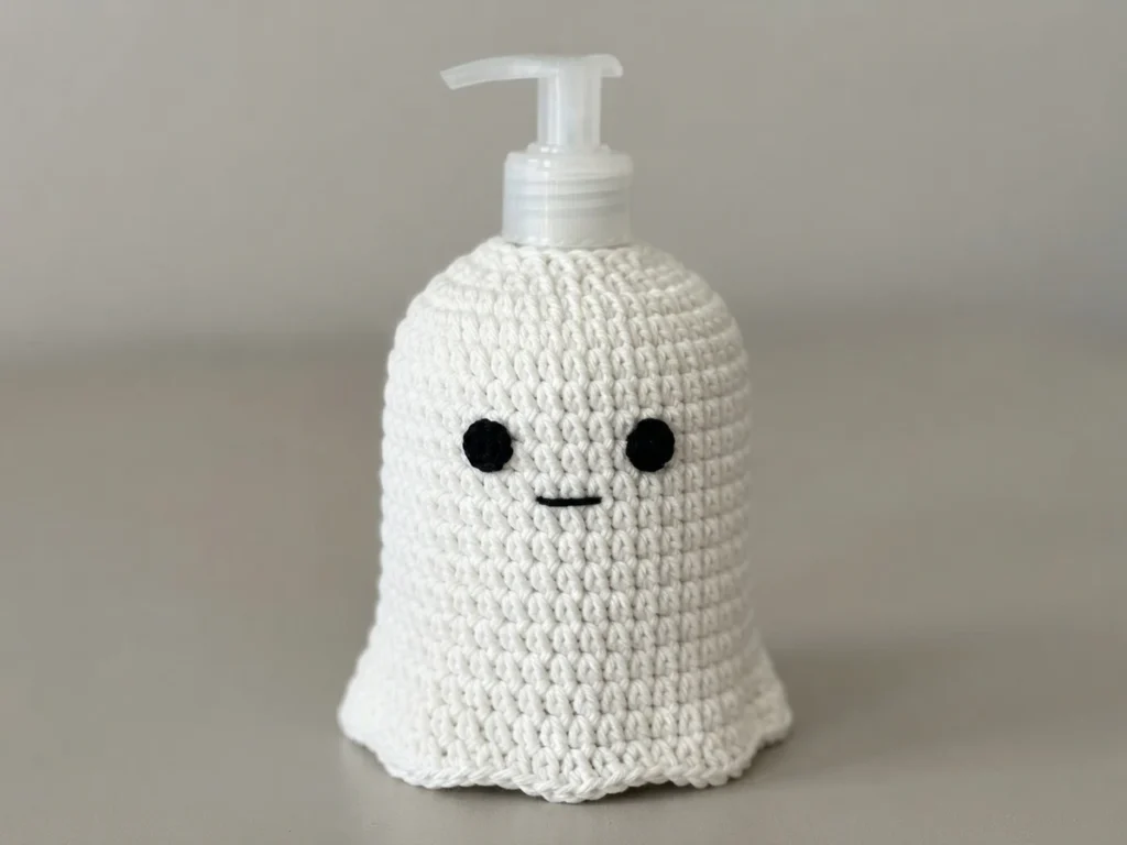 White crocheted ghost cover with two black eyes and a small black mouth, fitted over a clear plastic soap dispenser pump, photographed against a plain gray background.