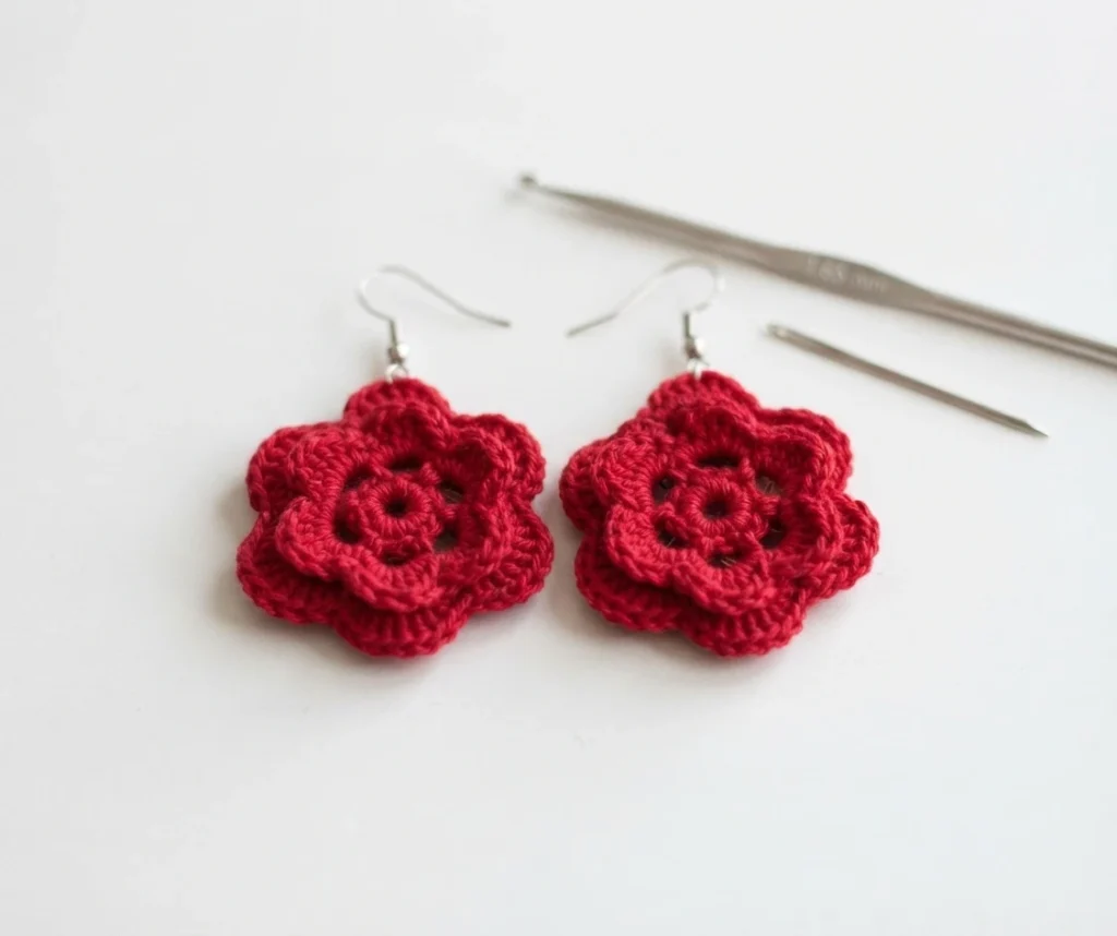 Pair of red layered crochet flower earrings with silver hooks lying on a white surface beside a steel crochet hook and a needle