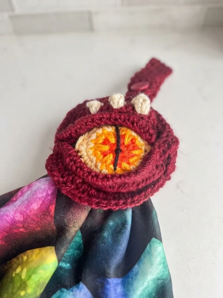 Close up of a burgundy crochet dragon eye towel topper with cream spikes, an orange and yellow eye and a multicolored dragon scale towel below