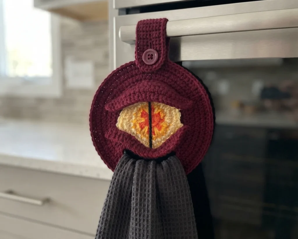 Burgundy crochet dragon eye towel topper with a yellow and orange eye hanging from an oven handle and holding a grey towel