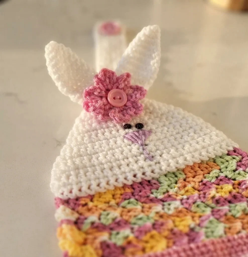 White crocheted bunny hanging towel with long ears, pink flower with pink button, small black eyes, and multicolor pink, yellow, green and orange body