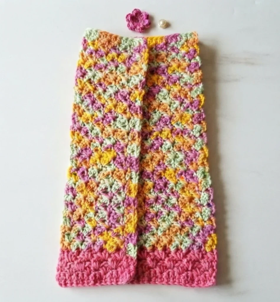 A crocheted towel in variegated pink, yellow, orange and green yarn with a pink bottom stripe, folded lengthwise with the sides overlapping, laid flat with a small pink crocheted flower and a round button placed above it.