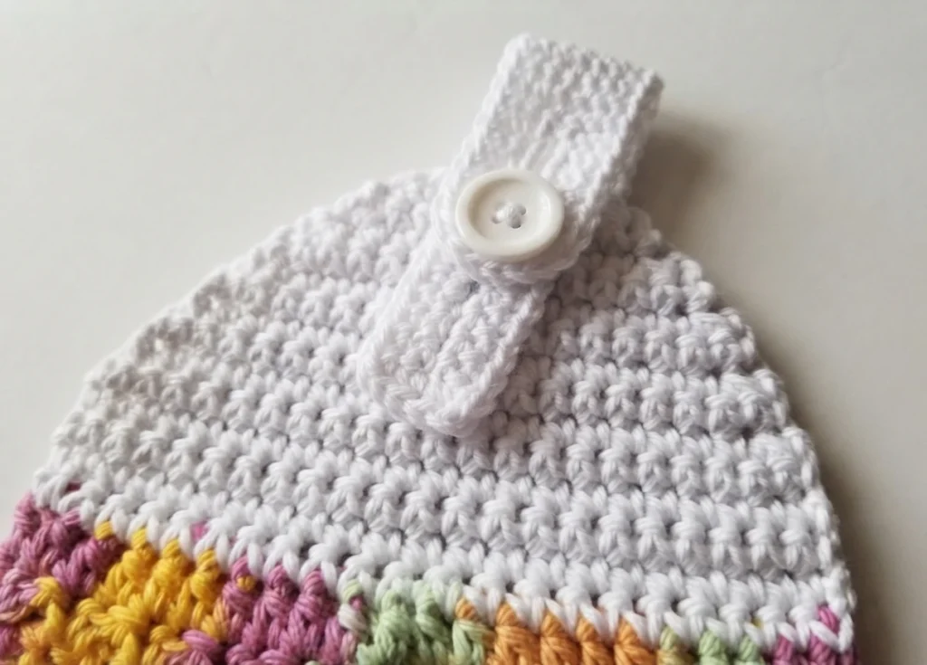 Close-up of the white crocheted top of a hanging towel with a buttoned hanging strap above a multicolor crocheted section