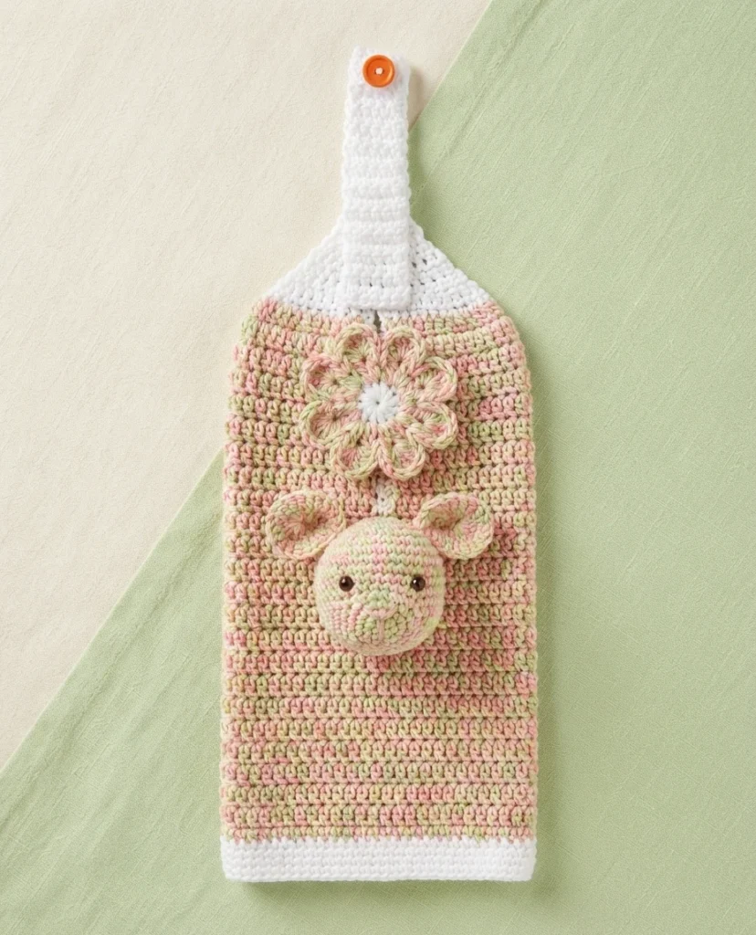 Crocheted hanging towel in pink and green variegated yarn with white top and bottom edges, orange button on the hanging strap, crocheted flower, and round bunny face applique with ears