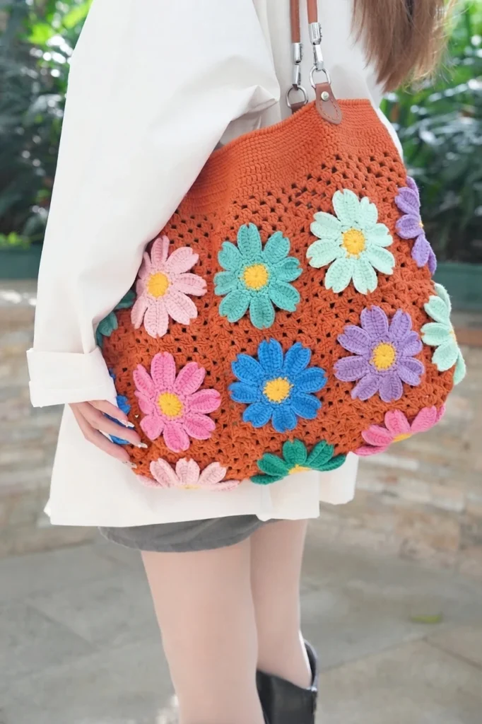 A woman in a white shirt carries a terracotta crochet tote covered with colorful daisy granny squares with yellow centers, on a brown leather shoulder strap.