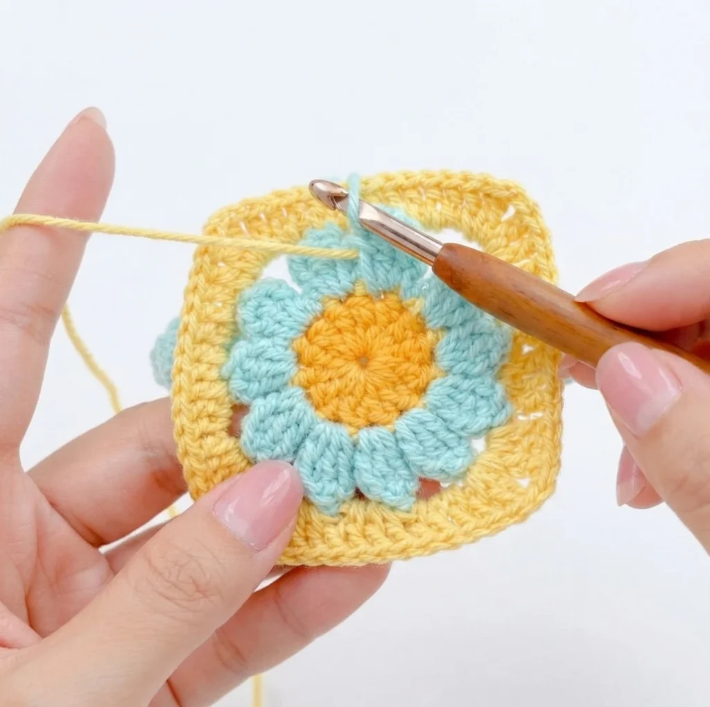 Hands holding a crochet daisy granny square with aqua petals, a yellow center and a yellow square border, a wooden-handled crochet hook and a strand of yellow yarn nearby.