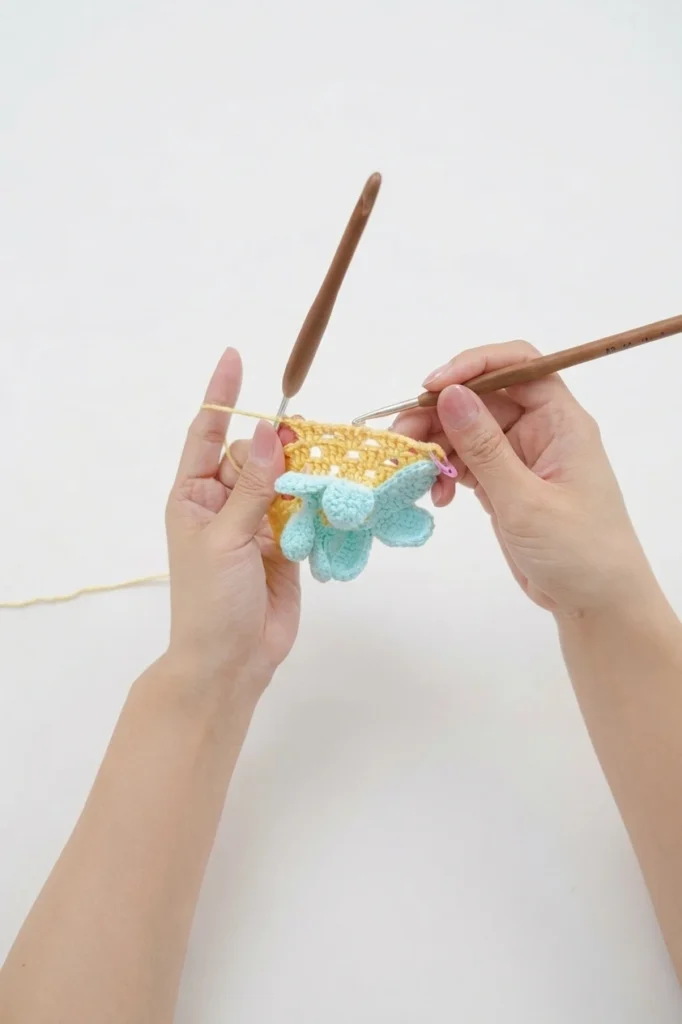Hands holding a small yellow crocheted mesh piece with a mint blue daisy flower, with two wooden crochet hooks on a white background.