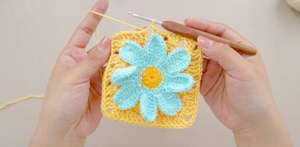 Hands holding a crocheted daisy granny square with turquoise petals and a yellow center on a yellow square, with a crochet hook inserted at the top.