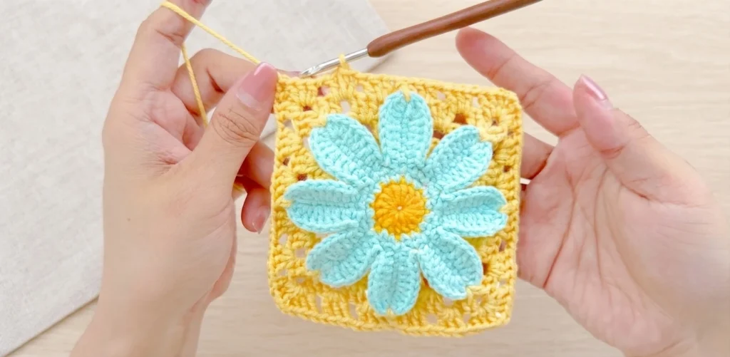 Hands holding a yellow crochet granny square with a light blue daisy and yellow center, with a crochet hook at the top edge