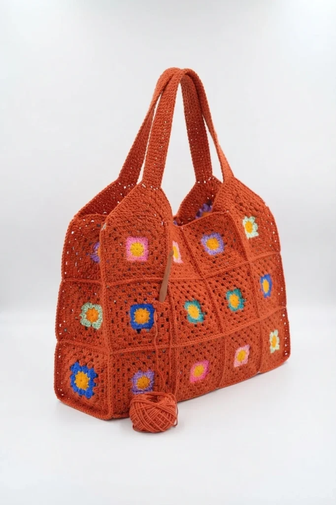 Terracotta crochet granny square tote bag with small colorful flower motifs, crocheted handles, and a ball of terracotta yarn