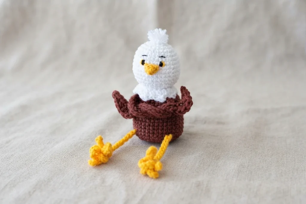 Crochet bald eagle amigurumi with white head, yellow beak and long yellow legs, brown body and raised wings on a beige fabric background